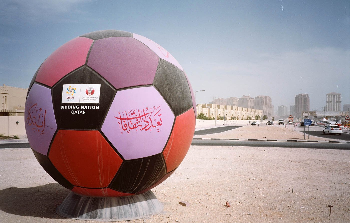L'immagine mostra un'enorme palla da calcio decorata, posizionata in un'area all'aperto. La palla presenta colori vivaci, inclusi viola e nero, e ha delle scritte decorative, probabilmente in arabo. Sul fronte, si vede un cartello con la scritta "BIDDING NATION" e il nome "QATAR", che indica il ruolo del Qatar come nazione candidata per un evento sportivo. Sullo sfondo, si intravedono edifici moderni e strade, in un contesto urbano. L'atmosfera è illuminata da una luce chiara, suggerendo una giornata di sole.