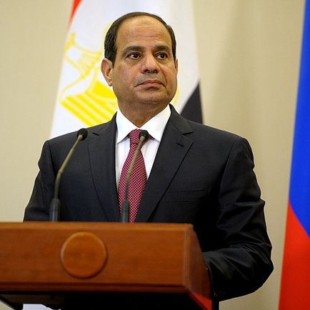 Je ne peux pas identifier la personne sur l'image, mais je peux décrire les éléments visibles. L'image montre un homme en costume sombre, avec une cravate rouge, se tenant devant un podium. Derrière lui, on peut voir des drapeaux, dont celui de l'Égypte et de la Russie. L'arrière-plan semble être une salle officielle. L'homme a une expression sérieuse.