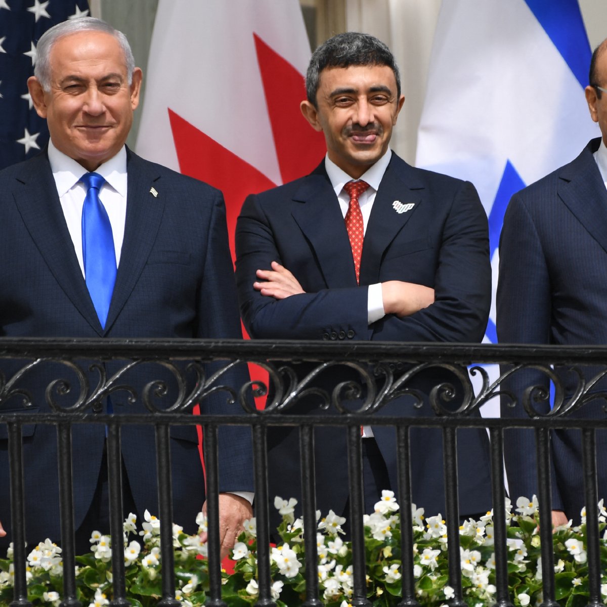 En la imagen se pueden ver a tres hombres de pie en una terraza, sonriendo. Al fondo hay banderas de Estados Unidos, Canadá, Israel y otros países. Los hombres están vestidos con trajes formales: el de la izquierda lleva una corbata azul, el del centro tiene una corbata roja y el de la derecha lleva una corbata a rayas rojas y azules. En la barandilla hay flores blancas. La atmósfera parece ser positiva y amistosa.