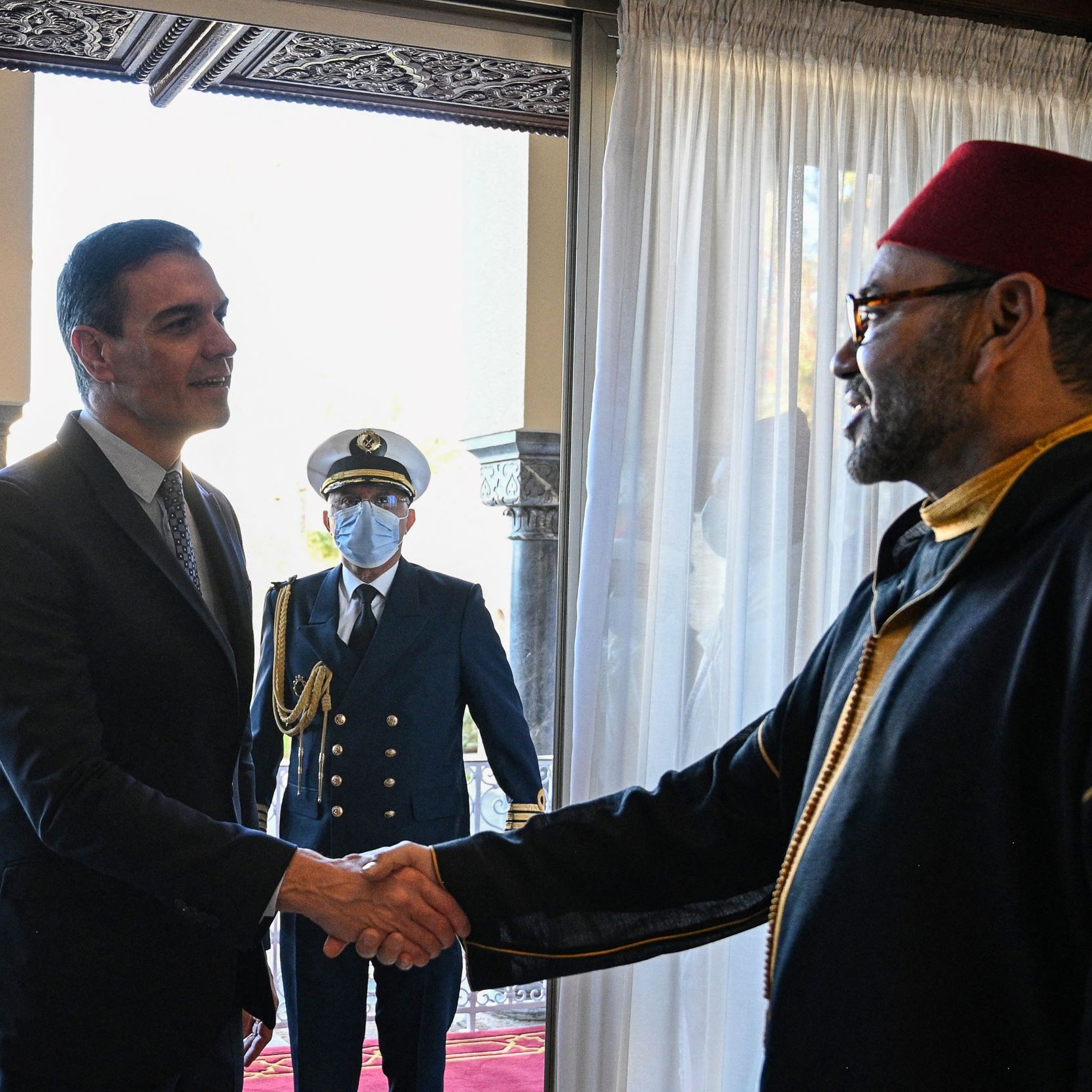 L'image montre deux hommes en train de se serrer la main dans un cadre intérieur élégant. L'un des hommes porte un costume, tandis que l'autre est habillé d'un habit traditionnel noir avec un chapeau rouge. En arrière-plan, un homme en uniforme militaire observe la scène. La lumière naturelle entre par une grande fenêtre avec des rideaux blancs, ajoutant une ambiance chaleureuse à la rencontre.
