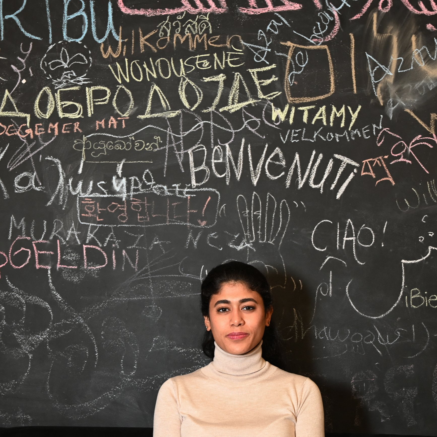 La imagen muestra a una joven posando frente a una pared de tiza oscura cubierta de múltiples palabras y frases escritas en diferentes idiomas. La mujer lleva un suéter claro y tiene el cabello recogido. La pared refleja un ambiente multicultural, con saludos y palabras de bienvenida en distintas lenguas.