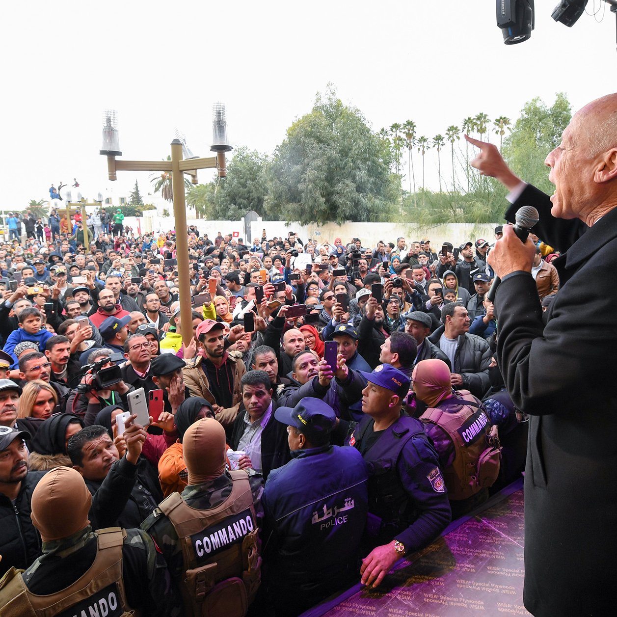 En la imagen se observa a un grupo numeroso de personas reunidas, probablemente en un evento o mitin. En el frente, hay un orador que está hablando con entusiasmo, utilizando un micrófono. Al fondo, se pueden ver elementos de seguridad, como un grupo de personas con chalecos que indican su función de protección o control del evento. El ambiente parece ser de gran expectación, y el público muestra interés en lo que se está diciendo. Además, hay un escenario o tarima donde se encuentra el orador. La escena transmite una sensación de participación activa de la gente.