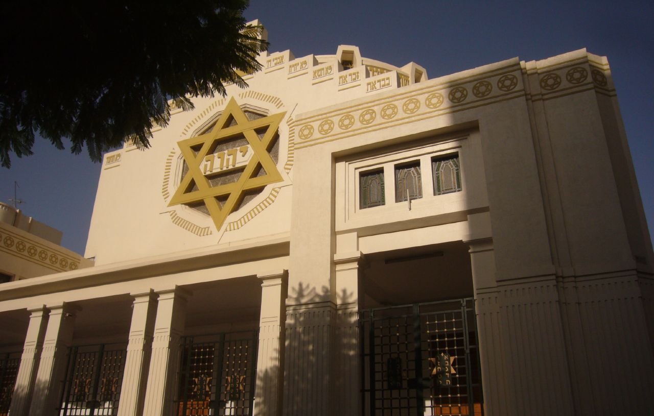 تظهر الصورة واجهة مبنى يمكن أن يكون معبدًا أو مركزًا ثقافيًا، يتميز بتصميم معماري فريد. الجدران الخارجية هي بألوان فاتحة، وهناك نجمة داود كبيرة مضاءة على الواجهة. يحيط بالمبنى أشجار خضراء، مما يضيف لمسة طبيعية. يبدو أن هناك نوافذ ذات تصميم زخرفي، مما يعطي المكان طابعًا مميزًا.