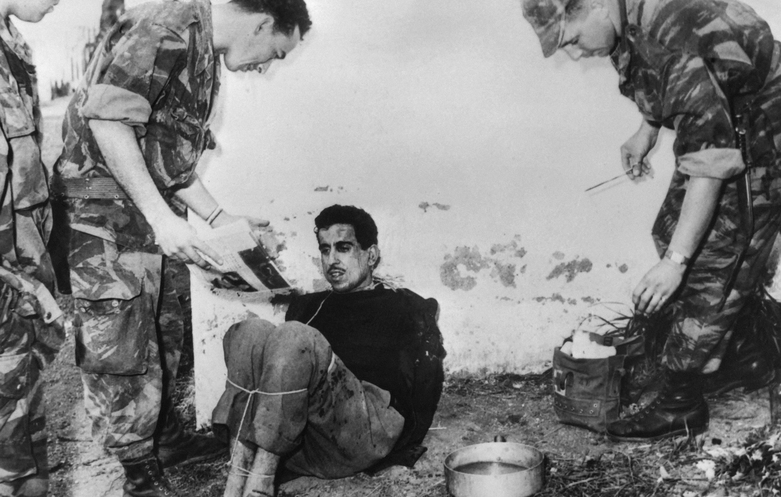La imagen muestra a un hombre sentado en el suelo, atado y aparentemente en una situación de interrogatorio o detención. Tiene una expresión de tensión en su rostro. Dos personas, vestidas con uniformes militares, están de pie a su alrededor. Una de ellas sostiene un documento y parece que está hablando con el detenido, mientras que la otra sostiene un utensilio, probablemente para tomar notas o hacer preguntas. El ambiente es austero y parece un lugar al aire libre, con una pared desgastada de fondo. La escena capta un momento de tensión y conflicto.