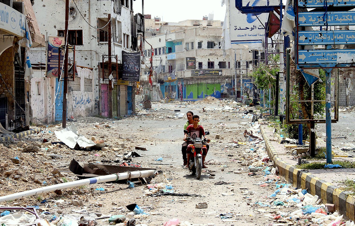 تظهر الصورة مشهداً لشارع في منطقة مهجورة أو متضررة، حيث تنتشر الفوضى والخراب. الشارع مليء بالقمامة والحطام، والمباني المحيطة تبدو متدهورة ومهجورة، مع بعض العلامات الإرشادية المتبقية. في وسط الصورة، يوجد شخصان يجلسان على دراجة نارية، مما يضيف لمسة من الحياة إلى المشهد القاتم. يبدو أن المكان يفتقر إلى الحركة والحيوية، مما يعكس حالة من الإهمال.