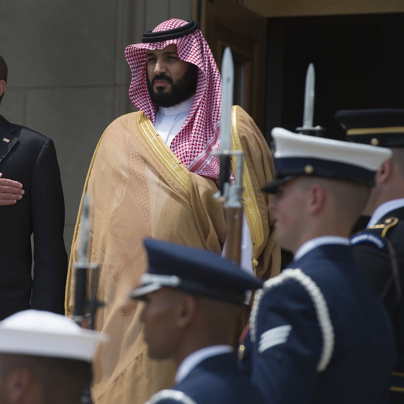 در این تصویر، دو مرد ایستاده‌اند. مرد سمت چپ با یک کت و شلوار تیره و پیراهن روشن دیده می‌شود و دستانش را بر روی سینه‌اش گذاشته است، که نشان‌دهنده احترام یا تقدیر است. مرد سمت راست در لباس رسمی عربی و با عمامه‌ای که به طور خاص مرتب شده، stands. در پس‌زمینه، گروهی از سربازان در یونیفورم‌های نظامی در حال ایستادن هستند. این صحنه احتمالاً به یک مراسم رسمی یا استقبال سیاسی مربوط می‌شود.