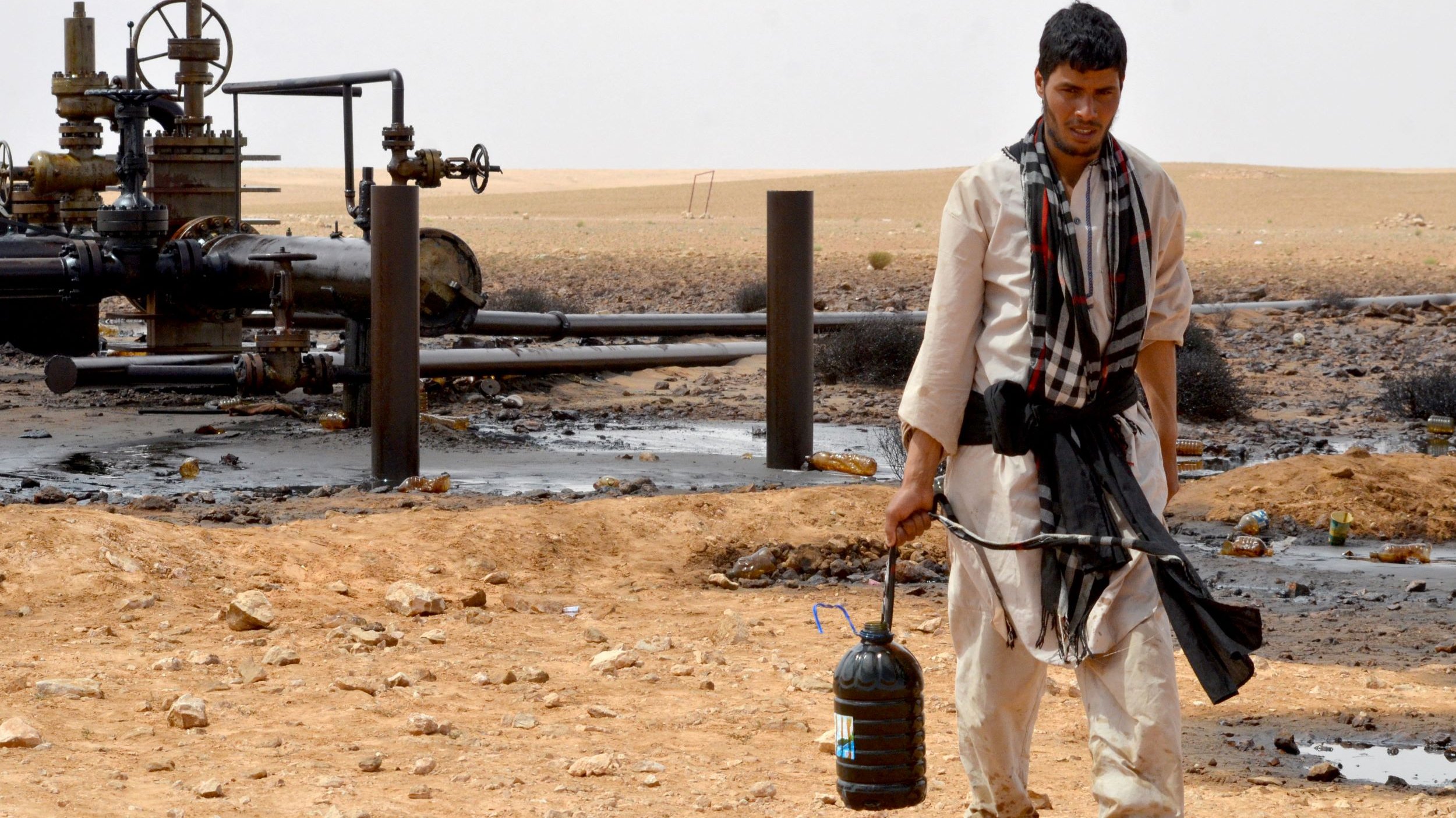 L'image montre un homme marchant à travers un paysage désertique. Il porte des vêtements traditionnels et a un seau noir dans une main. En arrière-plan, on peut voir des installations liées à l'industrie pétrolière, avec des tuyaux et des structures. Le sol est parsemé de pierres et il y a des traces de pétrole sur le terrain. L'environnement semble aride et désertique.
