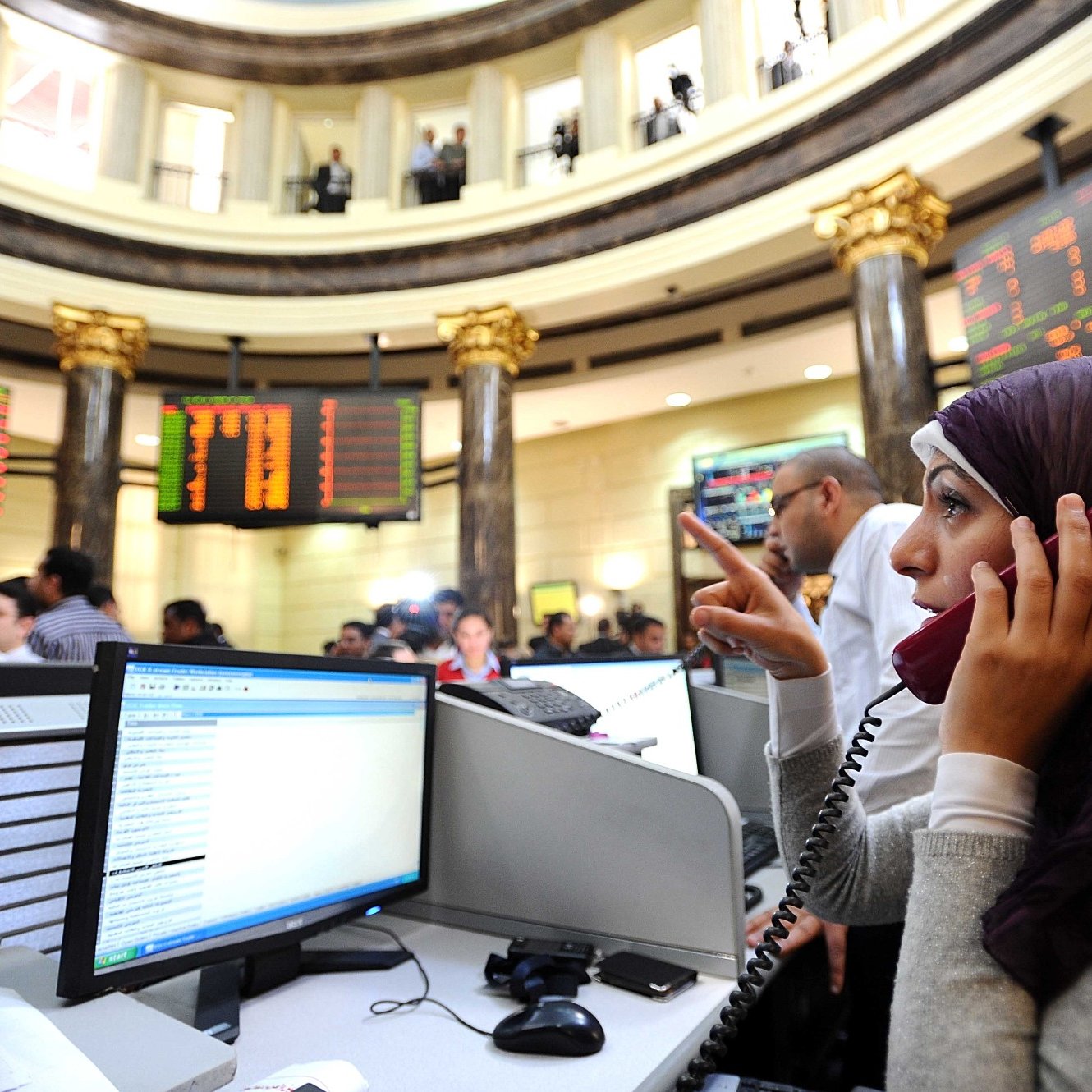 تظهر الصورة مشهداً من داخل سوق الأسهم، حيث يوجد عدد من الأشخاص يتعاملون مع شاشات تعرض الأسعار والبيانات المالية. في المقدمة، توجد امرأة ترتدي حجاباً، وهي تتحدث عبر الهاتف. يبدو أنها مشغولة بالعمل، حيث تشير بإحدى يديها. الخلفية تحتوي على عدة أشخاص آخرين وشاشات تعرض معلومات السوق. الأجواء توحي بالجدية والنشاط المالي.