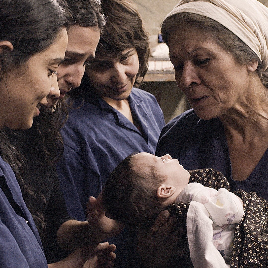 این تصویر گروهی از زنان را نشان می‌دهد که در یک محیط بسته و ساده قرار دارند. آن‌ها لباس‌های آبی رنگ به تن دارند و به یک نوزاد در آغوش یک زن مسن‌تر نگاه می‌کنند. زن مسن‌تر با محبت به نوزاد نگاه می‌کند و بقیه زنان نیز با چهره‌های شاد و پر از محبت به این لحظه توجه می‌کنند. فضای تصویر احساساتی از محبت و همبستگی را منتقل می‌کند.