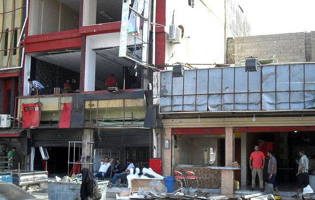 L'image montre un environnement urbain avec des bâtiments partiellement en ruines. On peut voir des façades de bâtiments endommagées, des débris éparpillés au sol et des personnes dans les environs, certaines visiblement en train de travailler sur la reconstruction. Il y a également des voitures garées à proximité. L'atmosphère semble être celle d'une zone en cours de rénovation ou de nettoyage après des dommages.