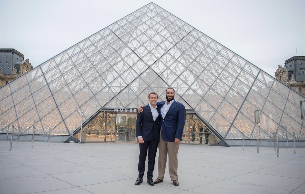 L'image montre deux hommes posant ensemble devant le Musée du Louvre, qui est célèbre pour sa pyramide en verre. Le bâtiment est moderne et impressionnant, contrastant avec l'architecture classique des ailes du musée. Les hommes sourient et portent des costumes formels, se tenant côte à côte, tandis que le ciel est légèrement nuageux, ajoutant une atmosphère calme à la scène.