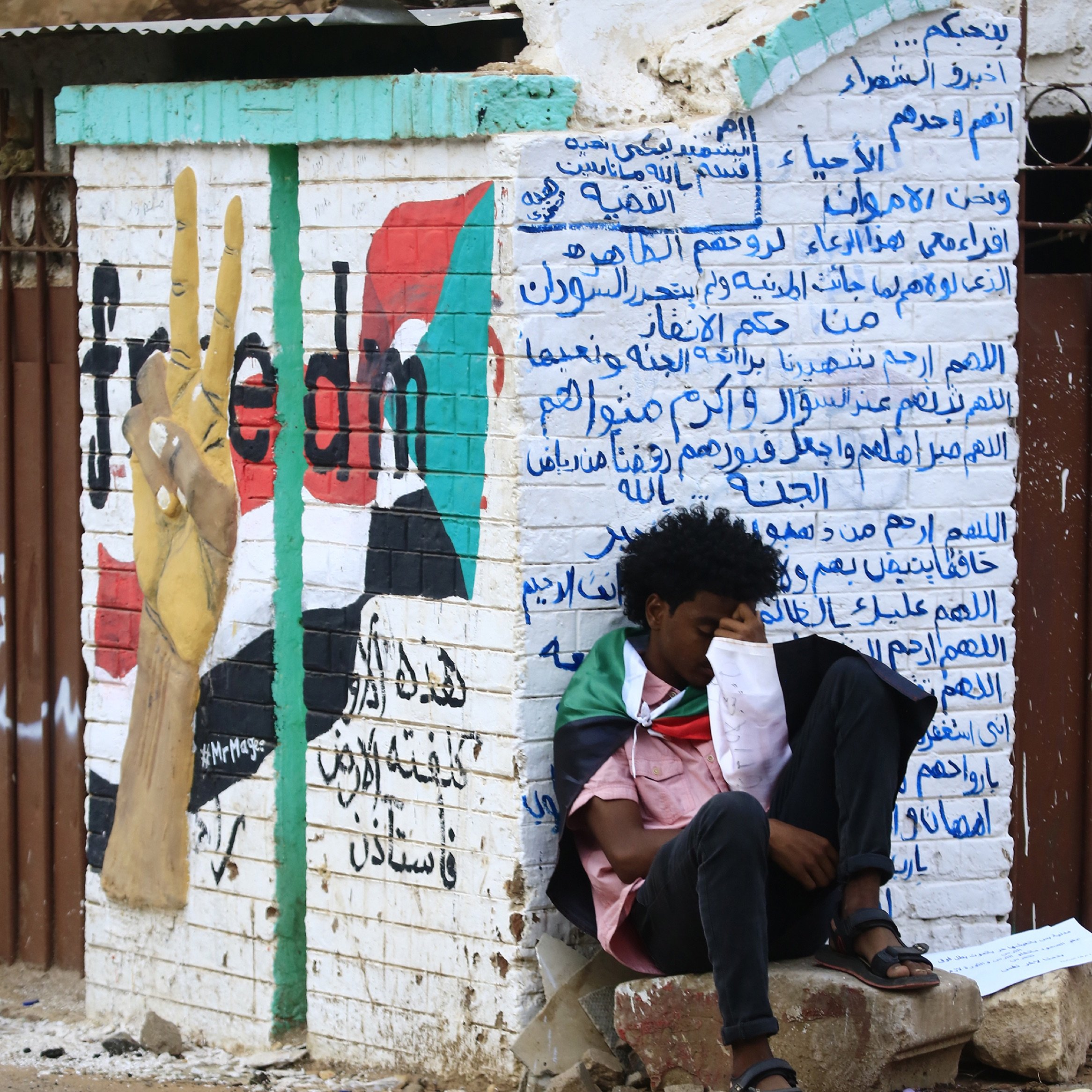 L'image montre un mur peint avec un symbole de paix, probablement représentant un mouvement pour la liberté. À gauche, il y a un graffiti d'une main levant deux doigts, symbolisant le mouvement pacifiste. Sur le mur, des inscriptions en arabe sont visibles, évoquant probablement des messages politiques ou sociaux. Au premier plan, un jeune homme est assis, enroulé dans un drapeau, semblant pensif ou mélancolique. L'atmosphère suggère un moment de réflexion sur des luttes pour la liberté.
