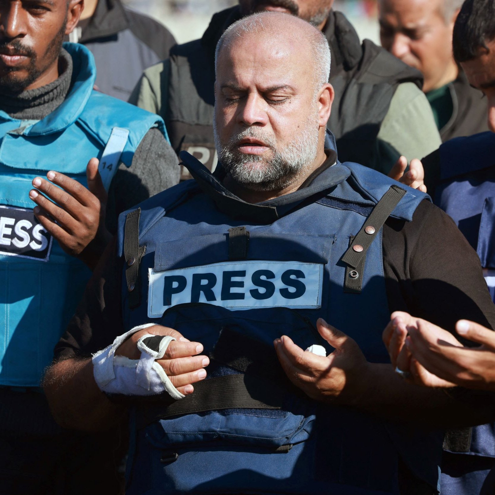 L'immagine mostra un gruppo di persone in piedi che sembrano partecipare a un momento di preghiera o riflessione. In primo piano, un uomo indossa un giubbotto con la scritta "PRESS" ed ha una fasciatura su un braccio. Gli altri membri del gruppo, che indossano abbigliamento simile, appare coinvolti nell'atto di pregare, con le mani sollevate. L'atmosfera sembra essere solenne e carica di emotività.