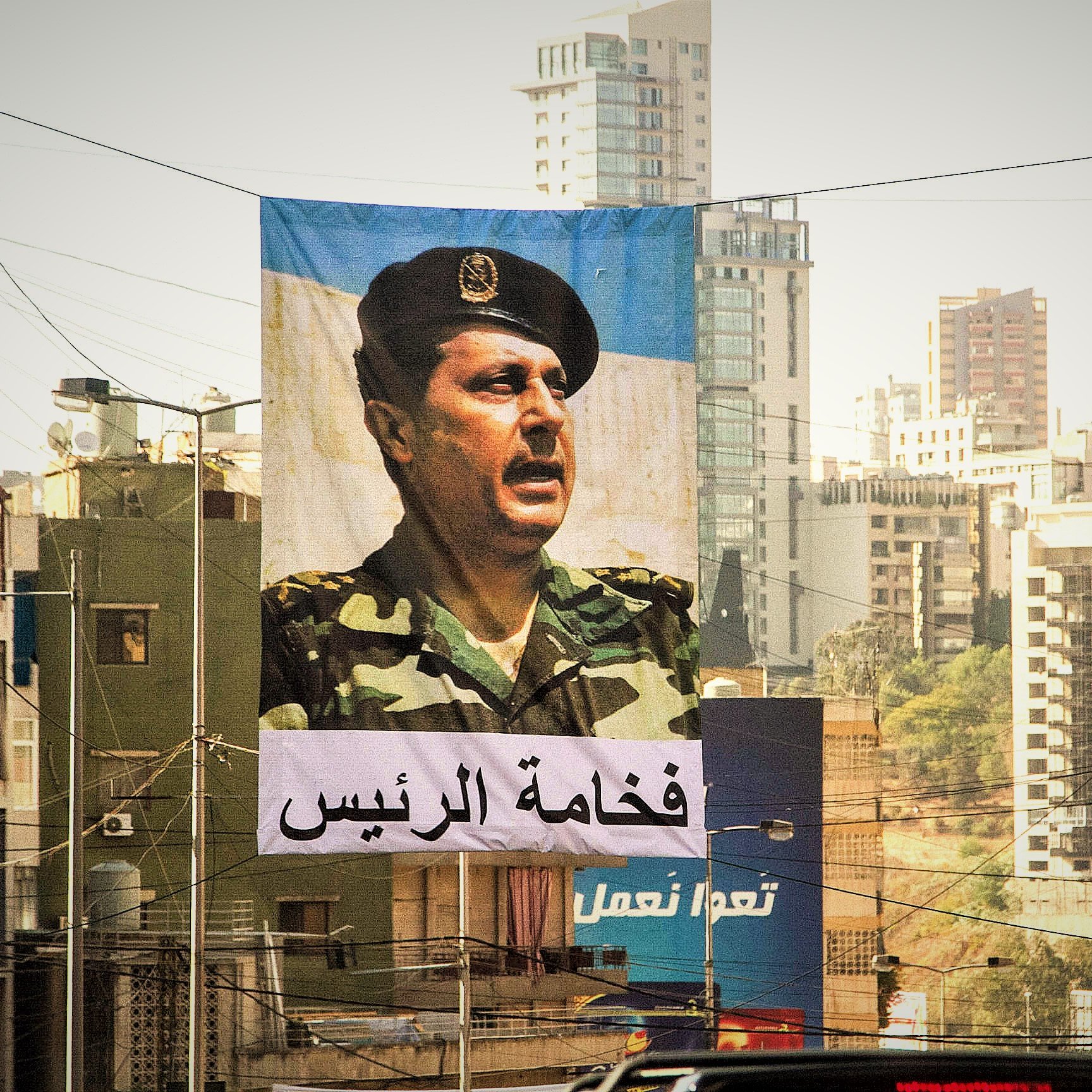 L'image montre une grande bannière suspendue dans une rue urbaine. Sur cette bannière, un portrait d'un homme en uniforme militaire est visible, face à un public. En dessous de l'image, il y a un texte en arabe qui semble faire référence à une autorité ou un leadership. En arrière-plan, on aperçoit des bâtiments modernes et d'autres structures urbaines typiques d'une ville. La lumière est douce, ce qui donne une atmosphère particulière à la scène.