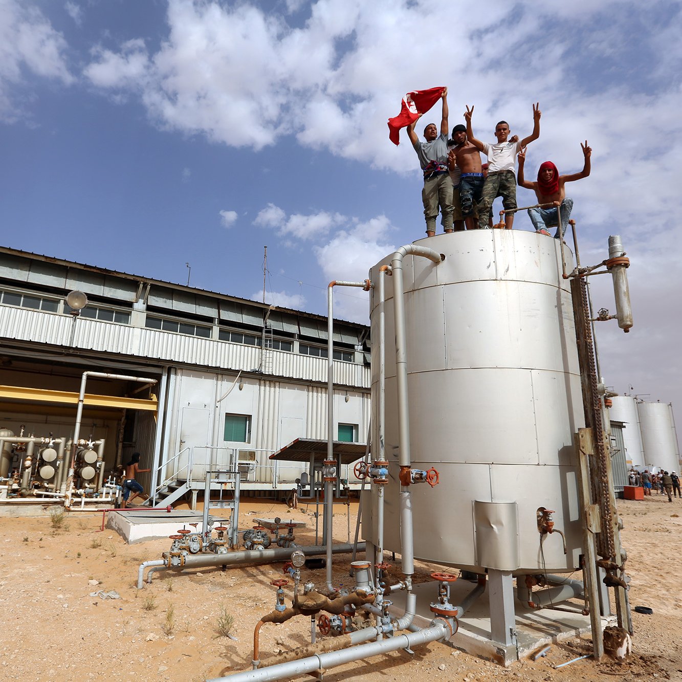 تظهر الصورة مجموعة من الأشخاص يقفون على قمة خزان معدني في منطقة صناعية، حيث يرفعون علمًا وينتشر خلفهم بعض المباني الصناعية. يبدو أنهم يحتفلون أو يعبرون عن فرحتهم، بينما يظهر في الخلف عدد قليل من الأشخاص الآخرين. السماء ملبدة بالغيوم، والأرض جافة. تعكس الصورة أجواء نشطة.