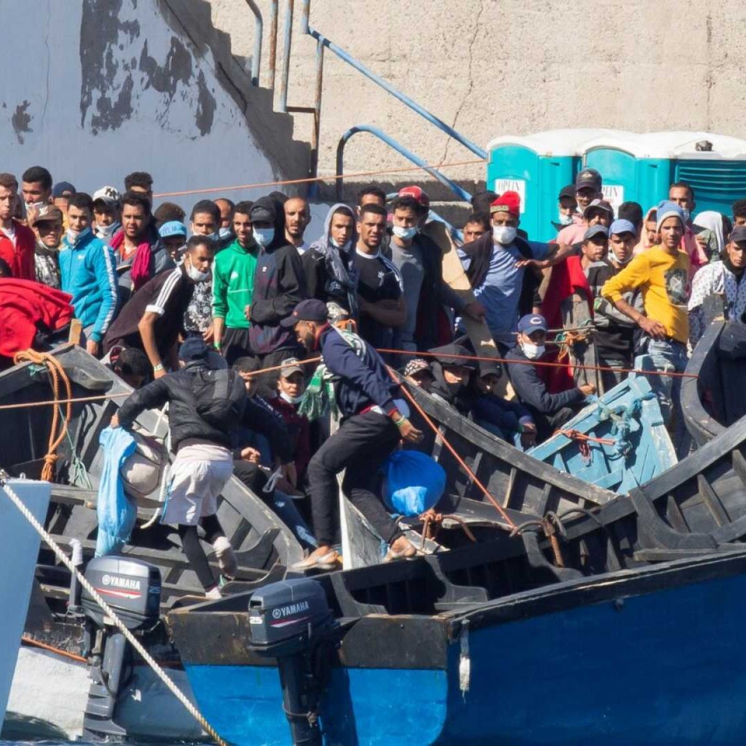 تظهر الصورة مجموعة من الناس يجتمعون حول قوارب في منطقة ميناء أو ساحل. يبدو أن هناك العديد من الأشخاص يتواجدون على القوارب، وبعضهم في وضعية استعداد للانطلاق. يرتدي البعض كمامات أو ملابس متنوعة، وهناك تجمع كبير من الناس يظهرون على جانب الميناء. الأجواء تعكس حالة من الحركة والنشاط، وربما تشير إلى عملية نقل أو هجرة.