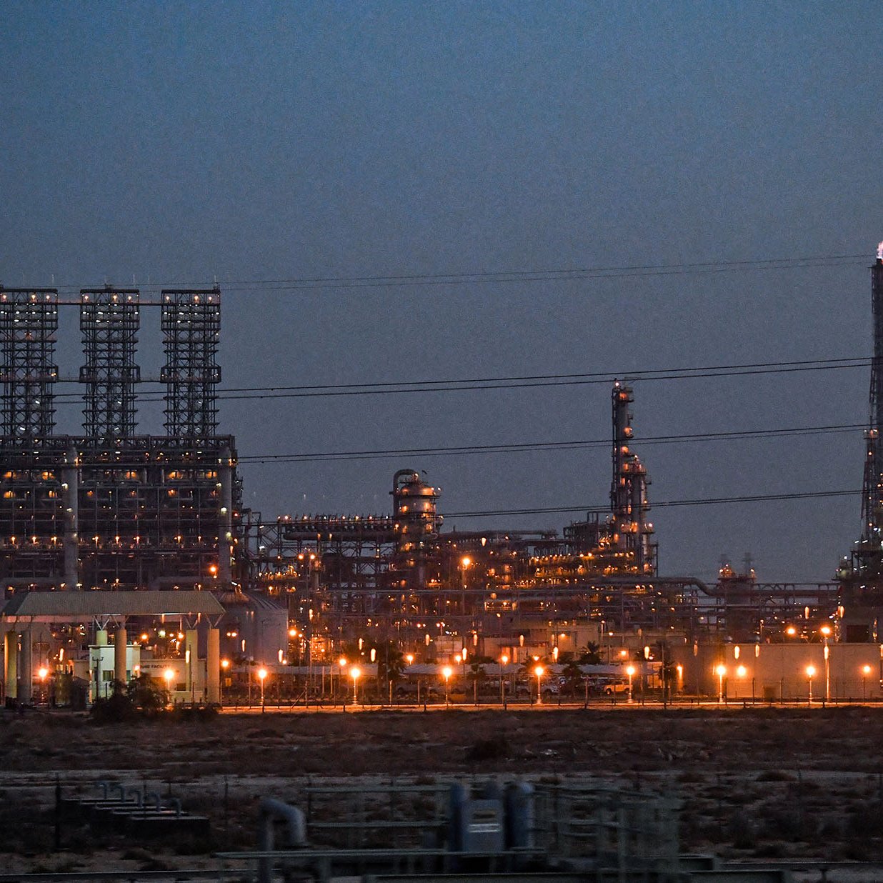 La imagen muestra una instalación industrial, probablemente una refinería o planta petroquímica, iluminada al anochecer. Se pueden ver numerosas estructuras metálicas altas, chimeneas y torres, con luces brillantes que resaltan en el entorno oscuro. Hay líneas eléctricas que cruzan la imagen y el paisaje es árido, lo que sugiere que la planta está ubicada en una zona desértica o poco vegetada. La atmósfera transmite una sensación de actividad industrial, con un enfoque en la tecnología y la producción.