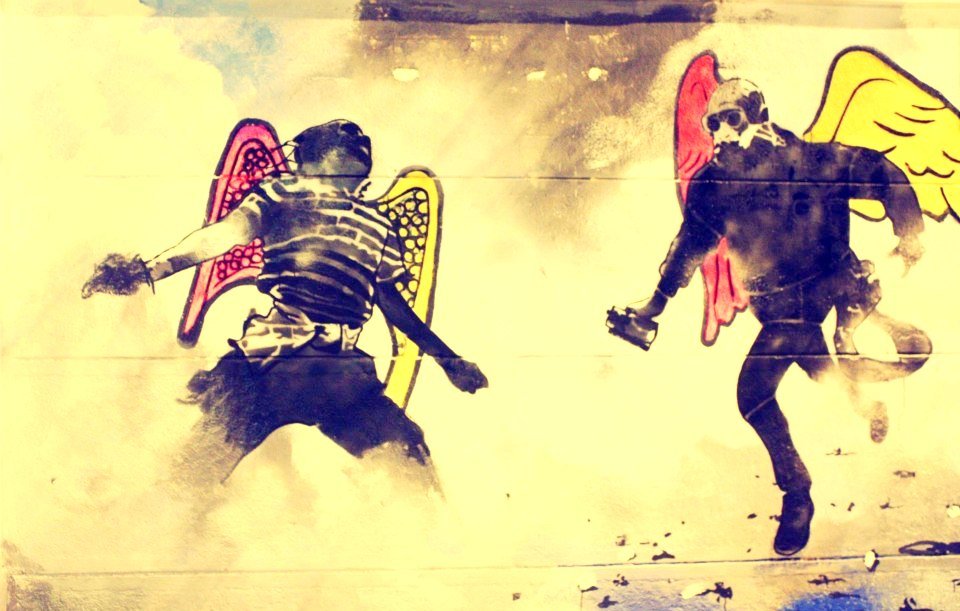 L'image présente deux figures humaines avec des ailes colorées, suggérant une représentation artistique et symbolique. L'une des figures porte un t-shirt rayé, tandis que l'autre est habillée en noir. Les ailes, d'un jaune lumineux pour l'une et d'un rouge pour l'autre, contrastent avec le fond plus sombre et nuageux, créant une atmosphère à la fois dynamique et énigmatique. Cette œuvre semble évoquer des thèmes de liberté et de conflit, donnant une impression de mouvement et d'énergie.