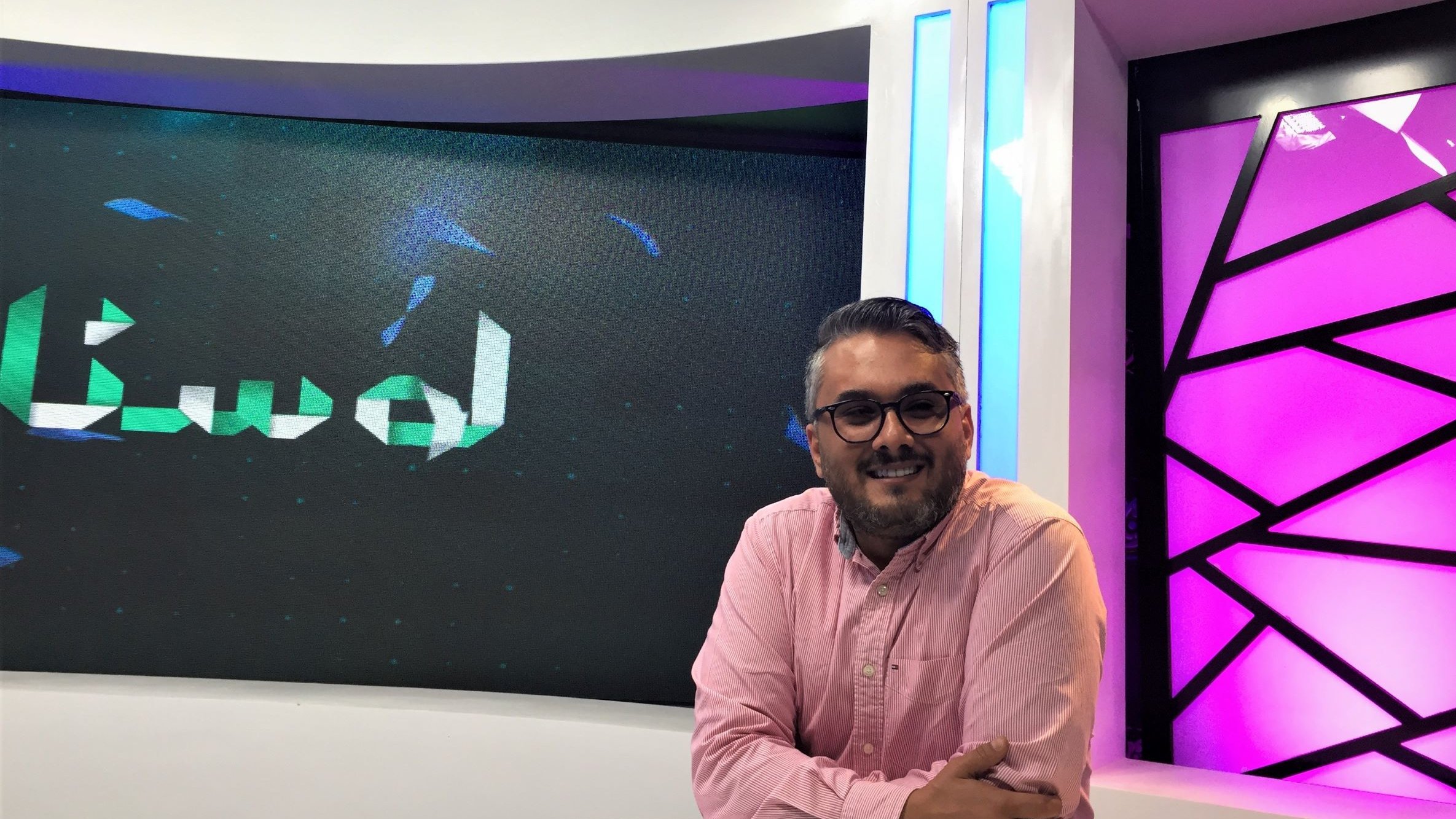 L'image montre un homme souriant assis devant un bureau dans un studio de télévision. Il porte une chemise rose et semble détendu. En arrière-plan, on peut voir un écran avec un motif abstrait et des lumières qui donnent une ambiance moderne au lieu. Le mot "لواح" est affiché sur l'écran, indiquant peut-être le titre d'une émission ou d'une rubrique. L'éclairage du studio est bien conçu, créant une atmosphère conviviale.