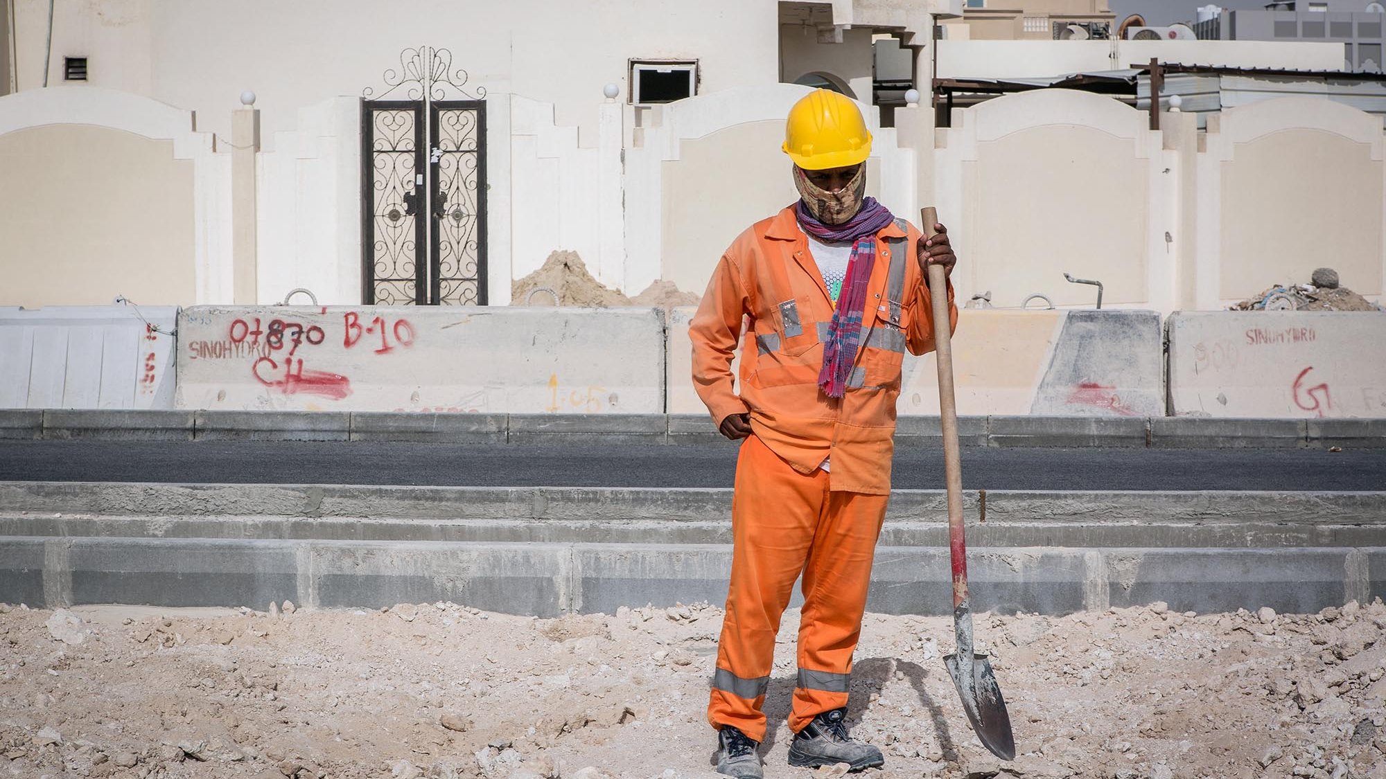 Nell'immagine si vede un lavoratore in un cantiere edile. Indossa una tuta arancione e un casco giallo per la sicurezza. Ha in mano una pala e sembra essere in pausa, osservando il terreno di fronte a lui. Sullo sfondo si possono notare alcune strutture e graffiti su un muro. L'ambiente appare polveroso e in fase di costruzione.