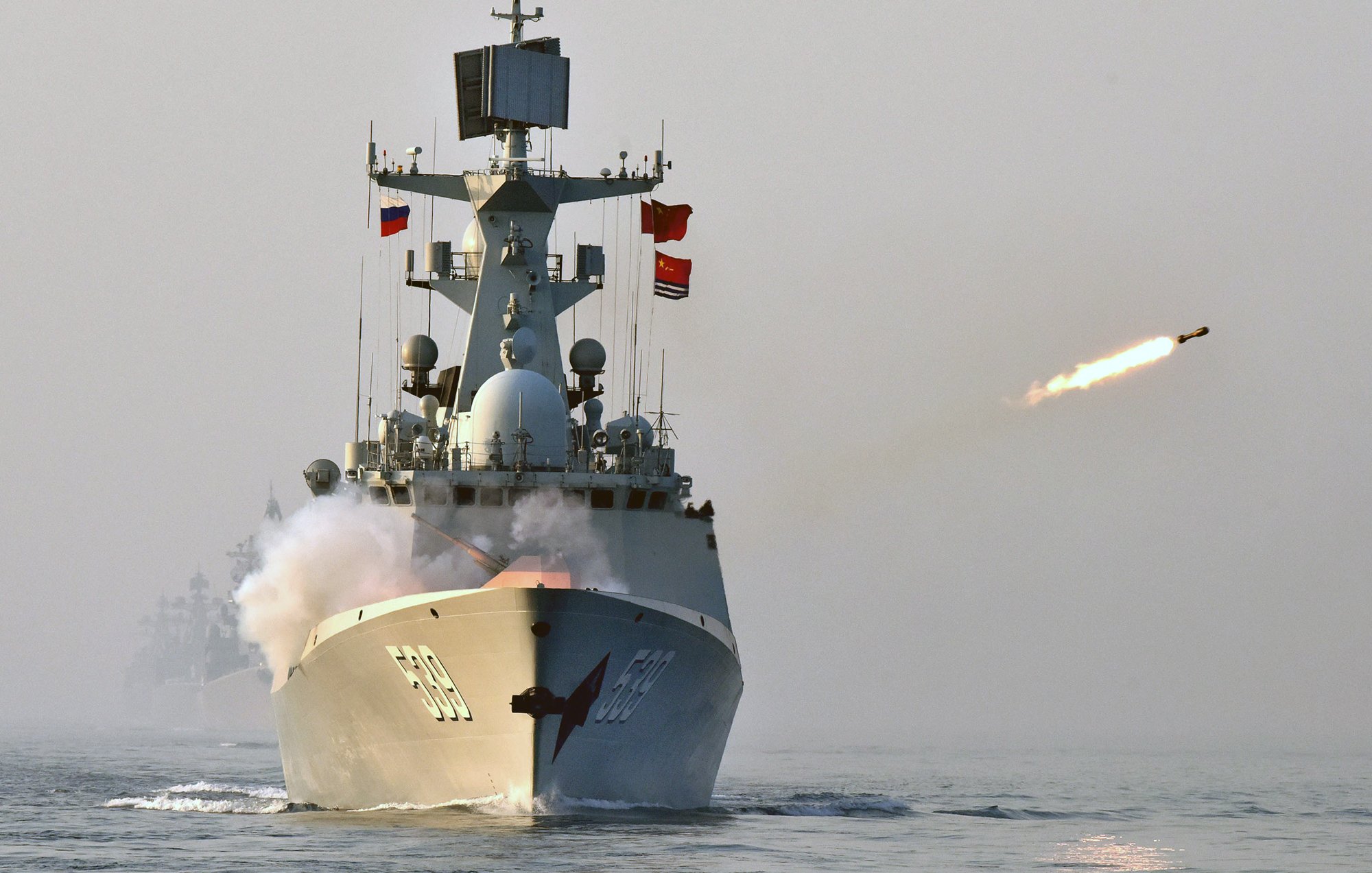 L'image montre un navire de guerre en mer, en train de tirer un missile. On peut voir de la fumée s'échapper du projetile, tandis que le bateau est orné de plusieurs drapeaux. L'atmosphère semble brumeuse, ce qui ajoute une certaine dramatisation à la scène. En arrière-plan, d'autres navires sont indistinctement visibles.