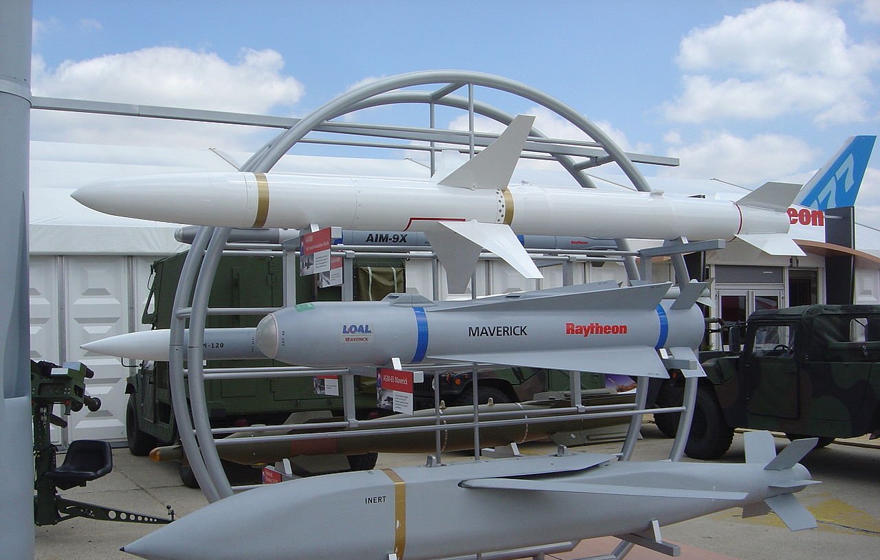 L'image montre une exposition de missiles sur un socle. On peut voir plusieurs types de missiles, dont un missile de croisière, un missile air-air et un missile guidé. Les missiles sont présentés sur un support circulaire, avec des informations en dessous de chacun d'eux. En arrière-plan, on aperçoit des véhicules militaires et un ciel partiellement nuageux. C'est une scène typique d'une foire ou d'une exposition de défense militaire.