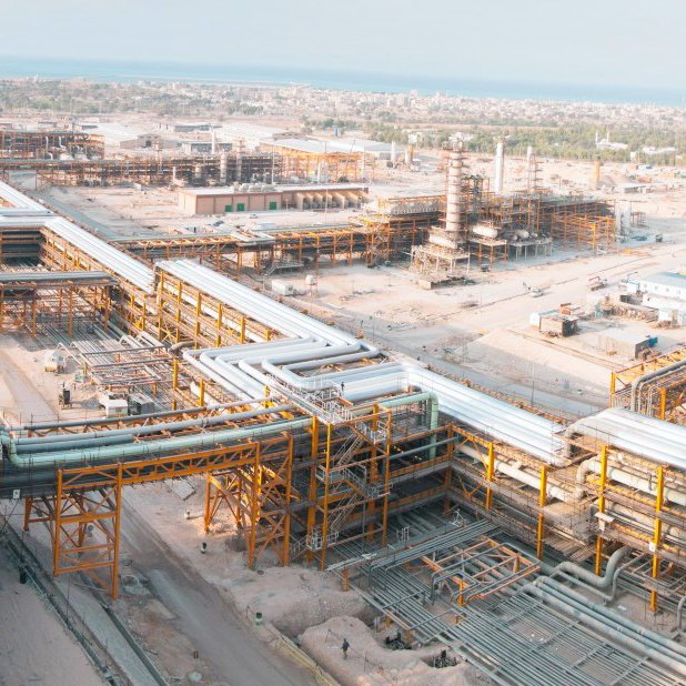 این تصویر یک مجتمع صنعتی بزرگ را نشان می‌دهد که شامل ساختارهای فلزی و لوله‌های گسترده است. در پس‌زمینه، تعدادی سازه بزرگ و تاسیسات به چشم می‌خورد. زمین اطراف به شکل خاکی و خشن است و در دوردست،‌ نوار آبی دریا نمایان است. این مکان به نظر می‌رسد که در حال فعالیت‌های صنعتی مانند پالایش نفت یا گاز باشد. فضا به طور کلی شلوغ و پر از تجهیزات و سازه‌ها است.