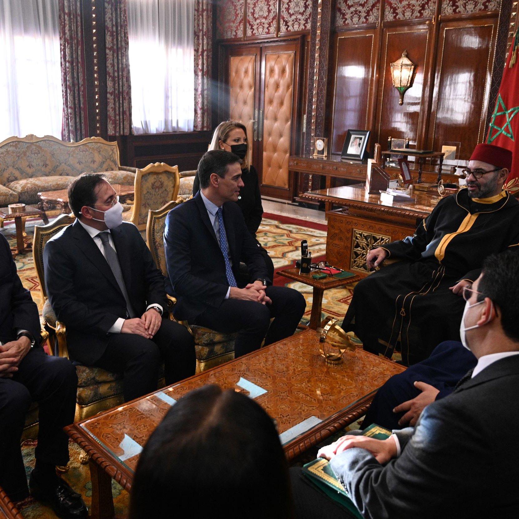 La imagen muestra una reunión en un ambiente formal y lujoso, posiblemente en un palacio. Hay varias personas sentadas alrededor de una mesa. Algunos de los asistentes llevan puesto un tapabocas, lo que sugiere que la reunión puede haber tenido lugar durante un periodo de preocupación por la salud pública, como la pandemia. En el centro de la escena, hay un individuo que parece ser una figura de autoridad, vestido con túnica y gorro tradicional. El interior está decorado con un estilo opulento, que incluye una alfombra elaborada y muebles de alta calidad. Las expresiones de los asistentes son serias, indicando la importancia del encuentro.