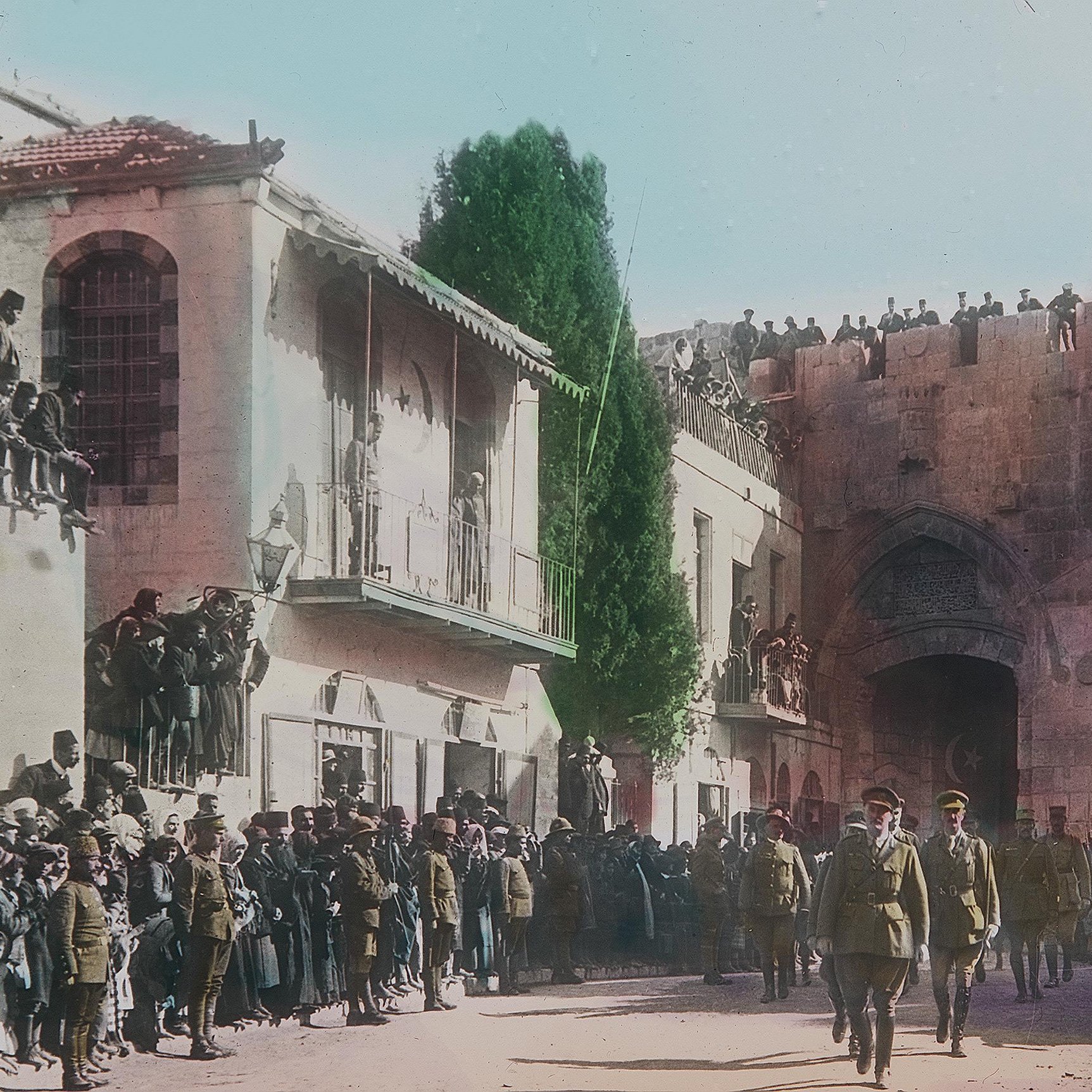 در این تصویر تاریخی، یک گروه از نظامیان در حال عبور از خیابانی در شهری با معماری قدیمی هستند. در دو طرف خیابان، جمعیتی زیادی ایستاده‌اند و به تماشای اتفاقات می‌پردازند. در پس‌زمینه، ساختمان‌هایی با طراحی خاص و دیواری بزرگ دیده می‌شوند. برخی از افراد در بالای دیوار ایستاده‌اند و نظاره‌گر هستند. فضای تصویر حسی از یک رویداد رسمی یا مراسم ویژه را القا می‌کند و رنگ‌آمیزی تصویر به آن جذابیت بیشتری بخشیده است.