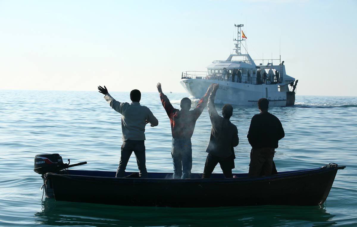 The image depicts four individuals in a small boat on calm waters, facing away from the camera. They are raising their arms, seemingly in a gesture of celebration or greeting. In the background, a larger boat is approaching, with several people visible on it. The scene is set against a clear sky, suggesting a peaceful day on the water.