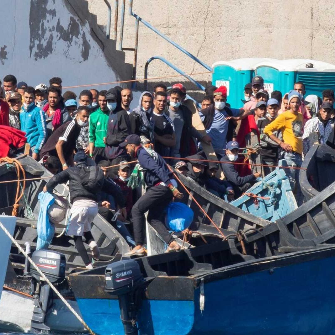 La imagen muestra un grupo de personas en un puerto o zona costera, donde hay varias embarcaciones. Muchas de las personas parecen estar en una o varias pequeñas embarcaciones de madera, mientras que otras están en la orilla o cerca de las embarcaciones. Se observa una multitud, con algunas personas vestidas en ropa casual, y parece que están en un momento de actividad o espera. Al fondo, hay estructuras de concreto y un baño portátil. La escena sugiere un ambiente de migración o búsqueda de oportunidades.