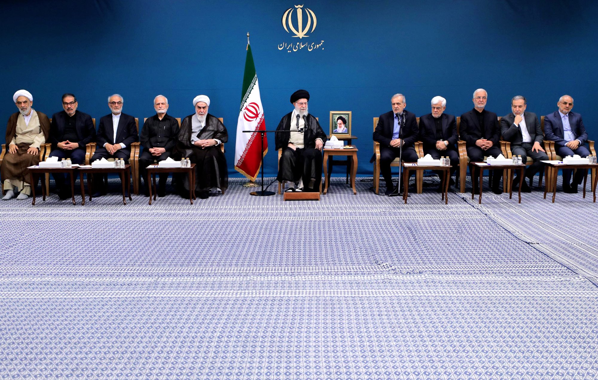 L'image montre un groupe de personnes assises en rangée sur un tapis, dans un environnement formel. En arrière-plan, il y a un drapeau iranien et un portrait d'une figure importante, probablement un leader. Les participants semblent être des hommes en costume sombre et en tenue religieuse, certains portant des turbans. Ils semblent engagés dans une discussion ou une réunion officielle. L'ambiance est solennelle et professionnelle.