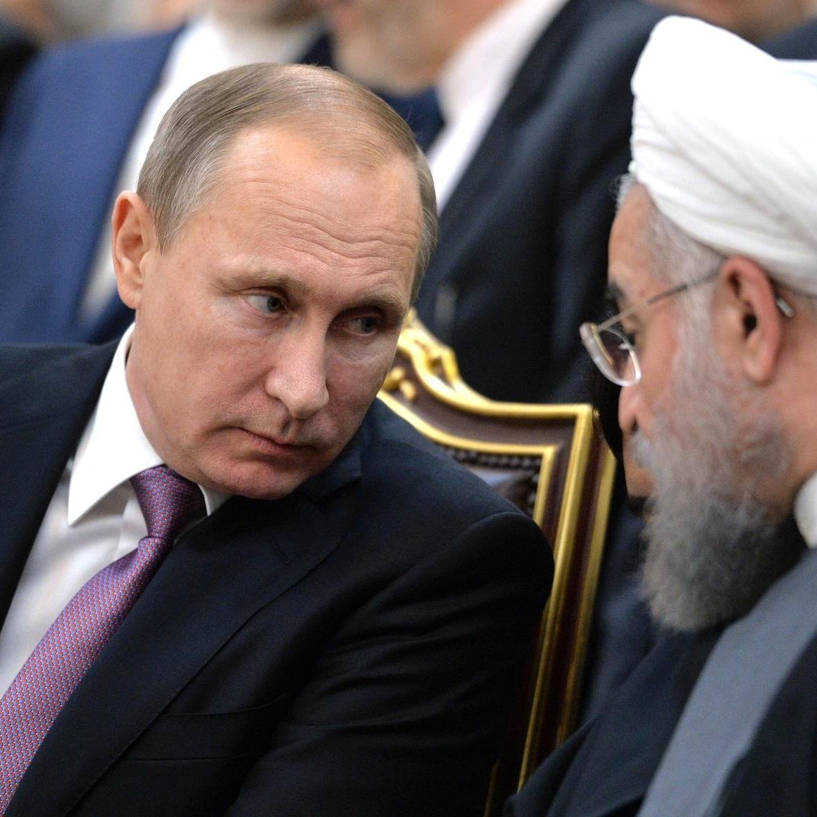 L'image montre deux hommes en costume, assis côte à côte lors d'un événement. L'un d'eux a les cheveux courts et une expression sérieuse, tandis que l'autre porte une barbe grise et semble en train de discuter. Ils sont entourés d'autres personnes floues en arrière-plan. Leur posture suggère une conversation attentive et concentrée.