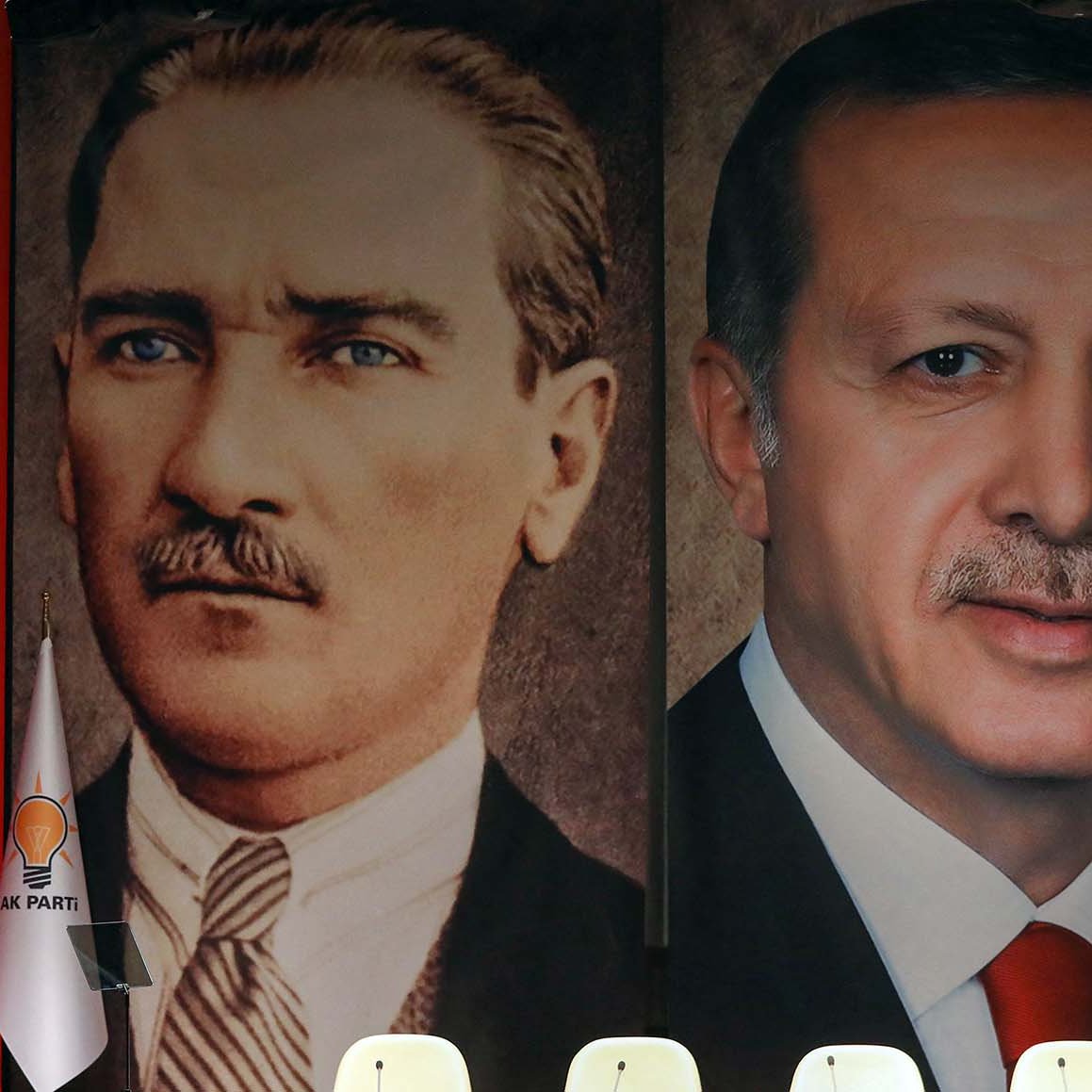 La imagen muestra a un orador frente a una gran bandera de Turquía, con retratos de dos figuras prominentes en la historia del país. A la izquierda, está el retrato de Mustafa Kemal Atatürk, el fundador de la República de Turquía, y a la derecha, el retrato del actual presidente, Recep Tayyip Erdoğan. El fondo es rojo, con símbolos nacionales y banderas que complementan el escenario, sugiriendo un evento político significativo.