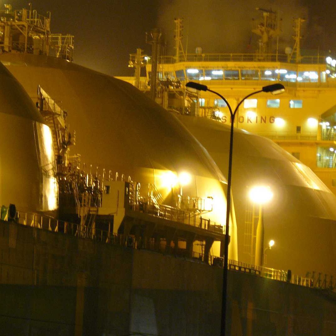 L'image montre une scène nocturne avec de grands réservoirs de stockage, probablement liés à l'industrie du gaz naturel. Les réservoirs ont des formes arrondies et sont éclairés par des lumières jaunes, créant une ambiance industrielle. En arrière-plan, on peut voir un navire, équipé de dispositifs de chargement ou de déchargement, suggérant une activité portuaire. La combinaison de l'illuminations et des structures métalliques donne une impression de travail intense et de technologie avancée.
