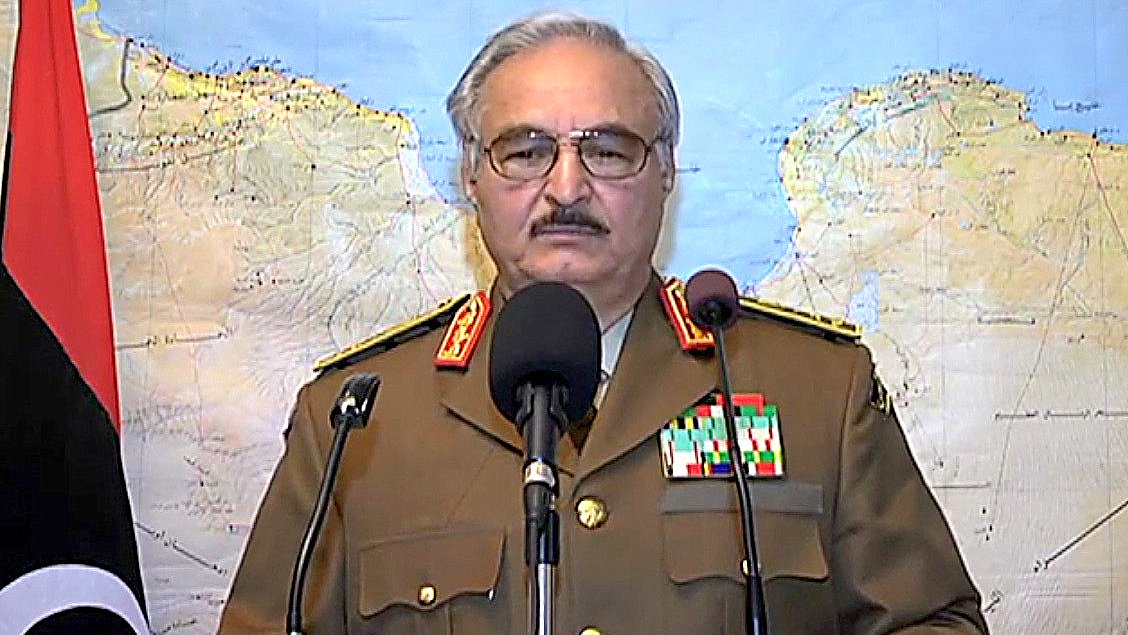 L'image montre un homme portant un uniforme militaire, avec plusieurs médailles et distinctions sur sa veste. Il se tient derrière un pupitre de conférence, devant une carte géographique en arrière-plan. L'homme semble s'adresser à un public ou à une caméra. On peut voir des drapeaux en arrière-plan, indiquant une situation officielle.