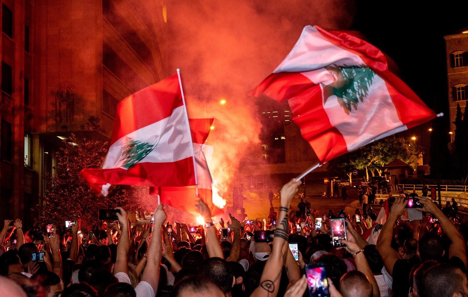 L'image montre une foule rassemblée, brandissant des drapeaux du Liban, dans une ambiance festive et animée. On peut voir des fumigènes rouges qui créent une atmosphère vibrante. De nombreuses personnes semblent capturer ce moment avec leurs téléphones. La scène se déroule probablement la nuit, avec des lumières urbaines en arrière-plan. L'engouement et la camaraderie parmi les participants sont palpables.
