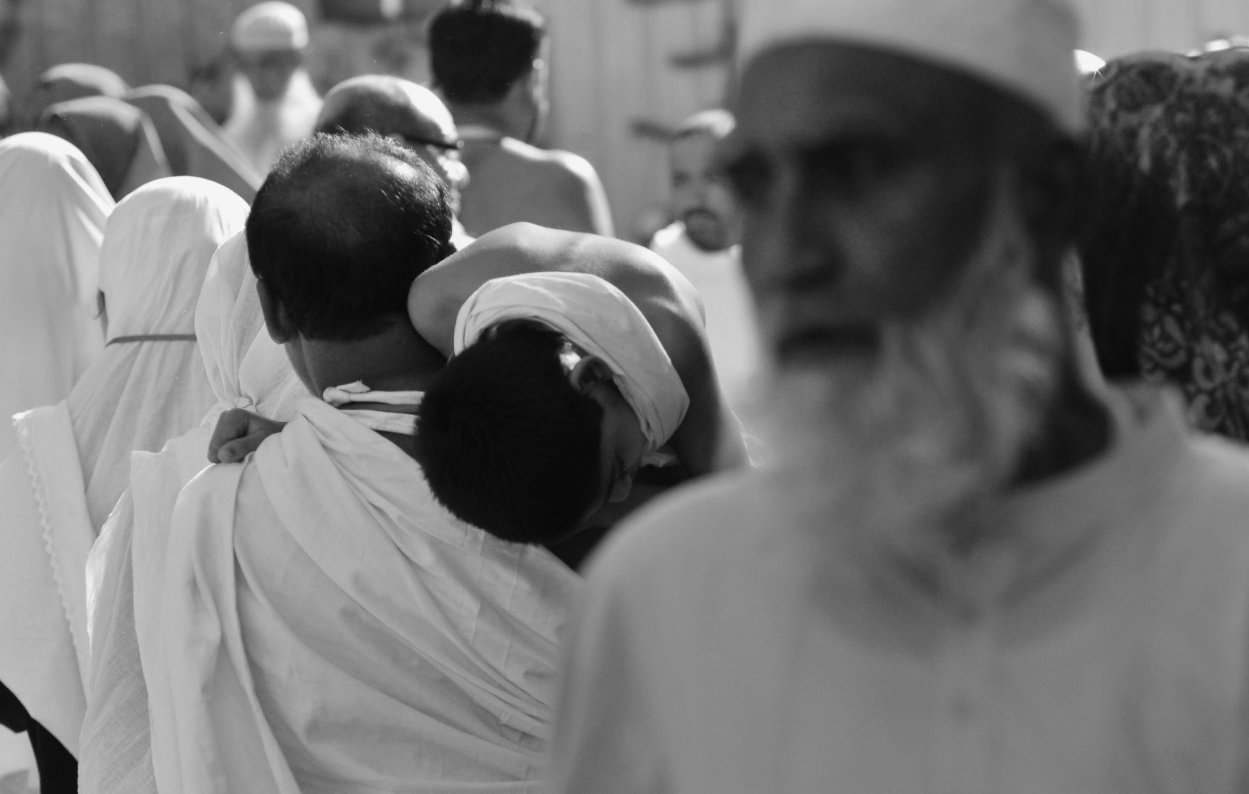 L'image montre une scène animée d'une foule, probablement lors d'un événement religieux. On peut voir un homme portant un enfant sur son dos, tandis que d'autres personnes sont visibles en arrière-plan, certaines portant des vêtements traditionnels. L'image est en noir et blanc, ce qui accentue l'atmosphère solennelle de l'événement. Les expressions des visages et les attitudes des personnes semblent refléter une ambiance de dévotion et de spiritualité.