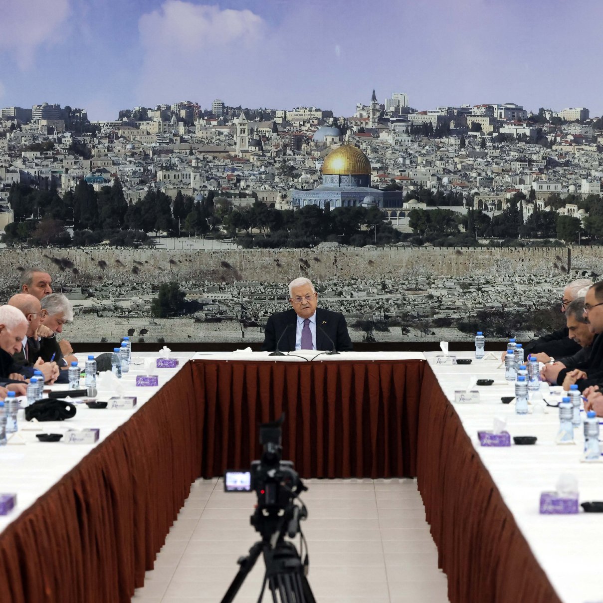 L'image montre une réunion se tenant autour d'une grande table ovale, avec plusieurs personnes assises de chaque côté. Au fond, il y a une grande image d'une ville, probablement Jérusalem, avec le dôme doré d'une mosquée bien visible. Les participants, qui semblent être des hommes âgés, portent des costumes sombres et affichent une expression sérieuse. Au centre de la table, une personne (probablement un chef ou un président) s'exprime pendant que les autres l'écoutent attentivement. Des bouteilles d'eau et des serviettes sont disposées sur la table. L'ambiance semble solennelle et attentive.