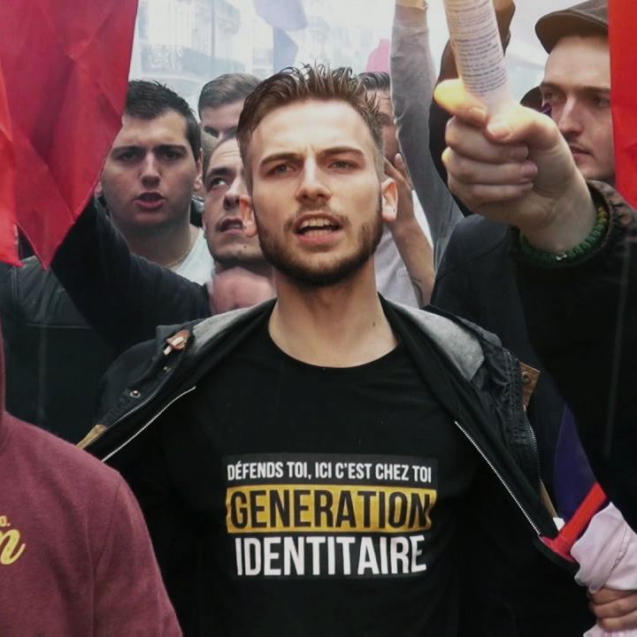 The image depicts a protest or demonstration scene. In the foreground, a young man stands prominently, wearing a black shirt with the text "GÉNÉRATION IDENTITAIRE" printed in yellow. He appears to be leading the group, holding a piece of paper in one hand and raising it in the air. Surrounding him are several other individuals, some of whom are holding red flags. The atmosphere seems charged and passionate, with many faces visible, indicative of a collective movement. The overall context suggests a rally focused on identity or nationalist themes.