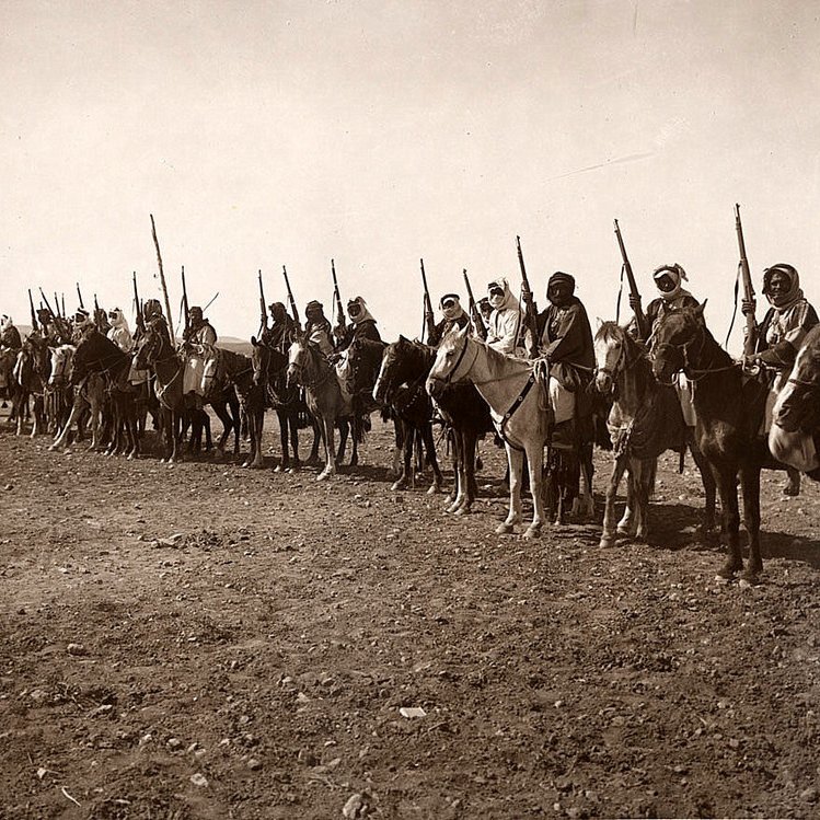 این تصویر نشان‌دهنده گروهی از سواران را در یک خط منظم است. آن‌ها بر روی اسب‌های خود نشسته‌اند و برخی از آن‌ها نیز نیزه یا سلاح‌هایی در دست دارند. لباس‌های آن‌ها سنتی به نظر می‌رسد و فضایی تاریخی را القا می‌کند. زمین زیر پای آن‌ها خشک و سنگی است و در پس‌زمینه، فضای باز و آسمان دیده می‌شود. این تصویر می‌تواند اشاره به یک دوره خاص از تاریخ یا مراسمی فرهنگی داشته باشد.