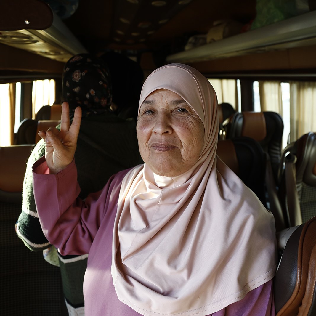The image shows an elderly woman wearing a light pink hijab, smiling and making a peace sign with her right hand. She is inside a vehicle that appears to be a bus, with other passengers visible in the background. The interior of the bus features comfortable seating and soft lighting, contributing to a warm atmosphere. The woman's expression conveys friendliness and warmth.