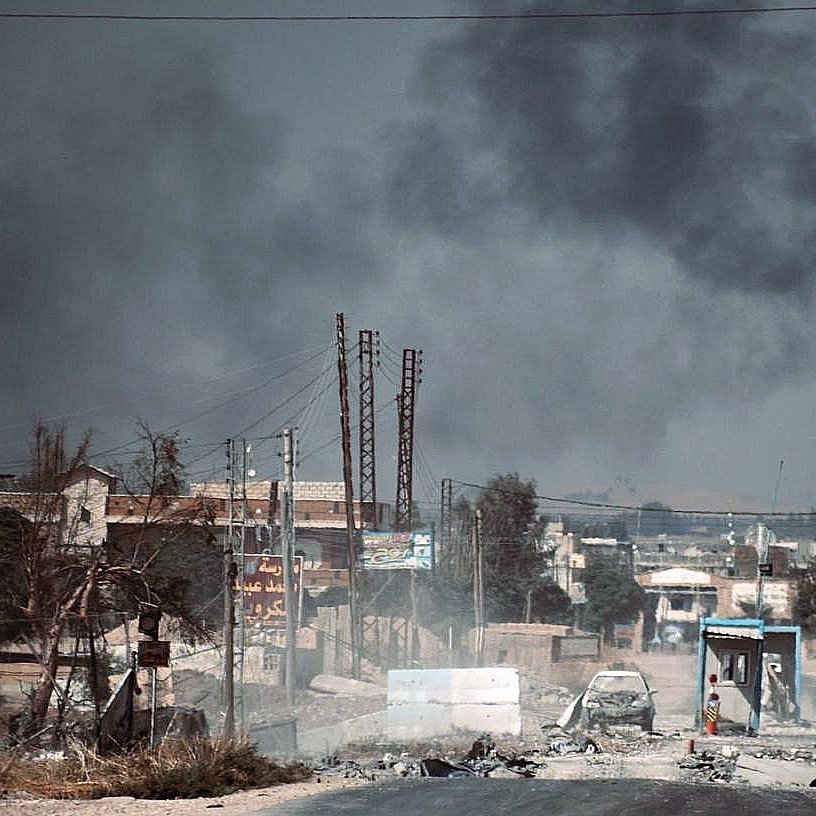تصویر نشان‌دهنده یک مسیر شهری در یک منطقه با وضعیت خراب است. در فضای پس‌زمینه، دود سیاهی از یک منبع آتش در حال بلند شدن است که نشان‌دهنده وقوع یک حادثه یا بحران است. ساختمان‌ها و سازه‌هایی در اطراف دیده می‌شوند که به نظر می‌رسد آسیب دیده‌اند. جاده‌ای که در تصویر وجود دارد، به سمت افق پیش می‌رود و نشانه‌هایی از تخریب و نابسامانی در آن به چشم می‌خورد. تعداد کمی از افراد در این صحنه حضور دارند که به نظر می‌رسد در حال عبور از این منطقه ناامن هستند. فضای کلی تصویر حس خفقان و ناپایداری را القا می‌کند.