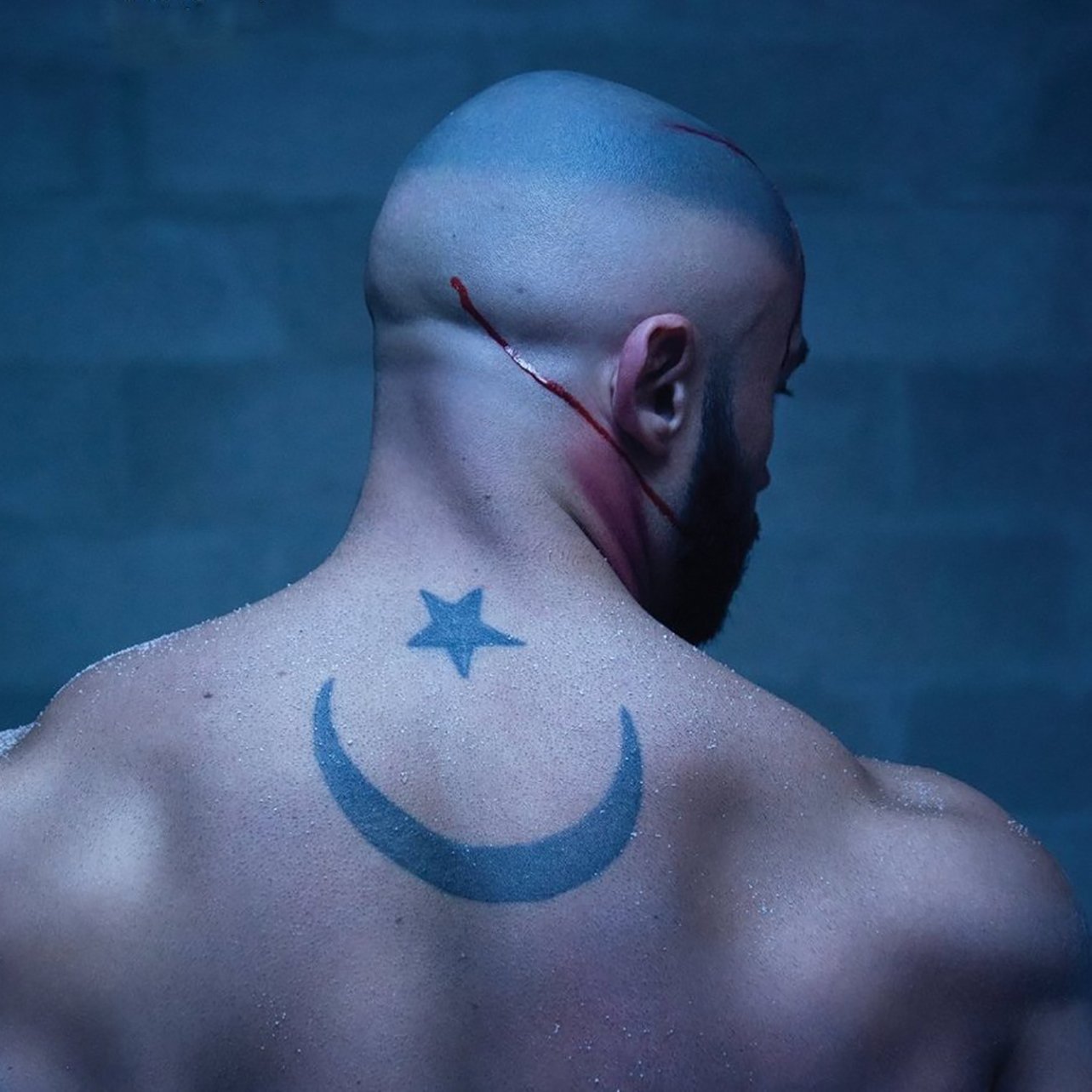 The image depicts a muscular, bald man from the back, showcasing a prominent tattoo on his back. The tattoo features a crescent moon and a star, which are often associated with various cultural and religious symbols. There are visible scars or cuts on his scalp and a streak of blood running down, suggesting a recent injury or intense situation. The background has a dark, moody atmosphere, possibly indicating a gritty or dramatic context.