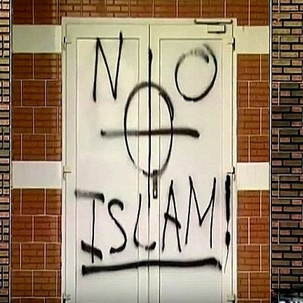 L'image montre une porte sur laquelle est peinte une inscription en spray noir. Le mot "NO" est écrit en haut à gauche, suivi d'un symbole ressemblant à une croix, et en dessous, le mot "ISLAM" est inscrit avec un point d'exclamation à la fin. Le fond de la porte et le mur environnant montrent des briques. L'ensemble de la scène évoque un message provocateur.