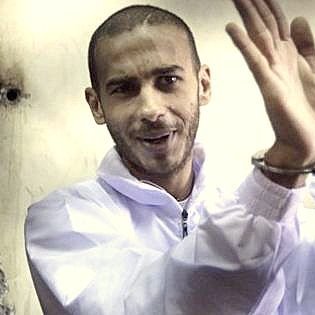 The image depicts a man in a white jumpsuit, appearing to smile or show a light-hearted expression. He is wearing handcuffs and seems to be gesturing with his hands, possibly in a playful or confident manner. The background appears to be a dimly lit space, which adds to the atmosphere of the scene.