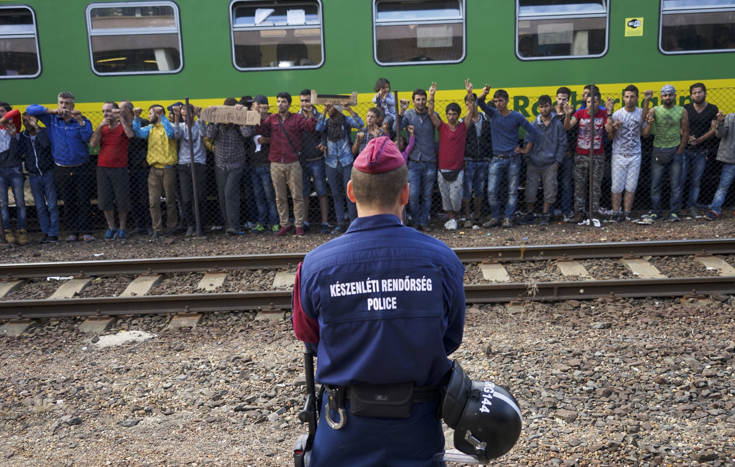 The image shows a scene at a train station where a police officer is facing a group of individuals. The officer is dressed in a uniform with the word "POLICE" clearly visible on the back. In front of the officer, there is a crowd of people, some holding signs, who appear to be expressing their discontent or making a statement. The background features a green train, and there are railway tracks visible, indicating that this is a transportation hub. The atmosphere seems tense, with the officer standing guard as the crowd raises their hands, possibly in protest or to seek attention.