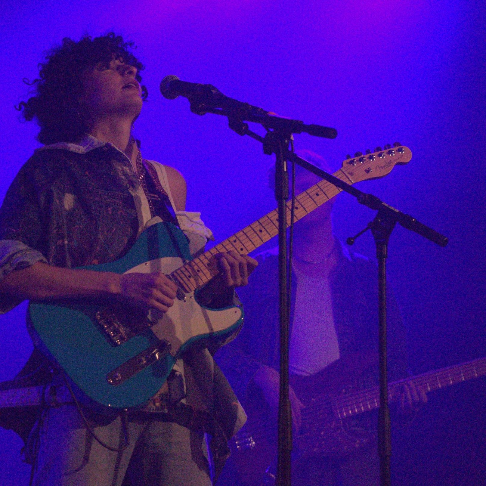 L'immagine mostra un musicista sul palco, intento a suonare una chitarra elettrica di colore azzurro. Ha i capelli ricci e indossa una camicia ampia con un look casual. Dietro di lui si intravedono altri musicisti e luci bluastre che creano un'atmosfera suggestiva. L'artista sembrano concentrato e coinvolto nella sua performance.