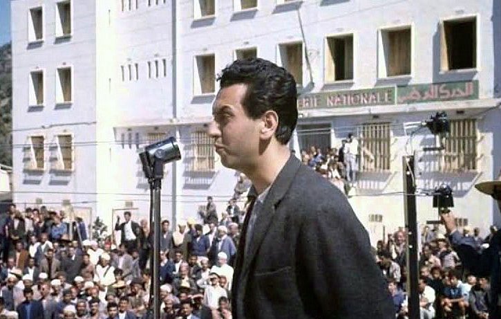 L'image montre un homme se tenant derrière un microphone, semblant s'adresser à une grande foule rassemblée devant lui. L'arrière-plan présente un bâtiment de style architectural simple avec plusieurs fenêtres, possiblement dans un contexte historique ou politique. Les membres de la foule semblent attentifs à son discours. L'éclairage naturel donne une ambiance claire à la scène, suggérant qu'elle pourrait avoir été prise en plein jour.