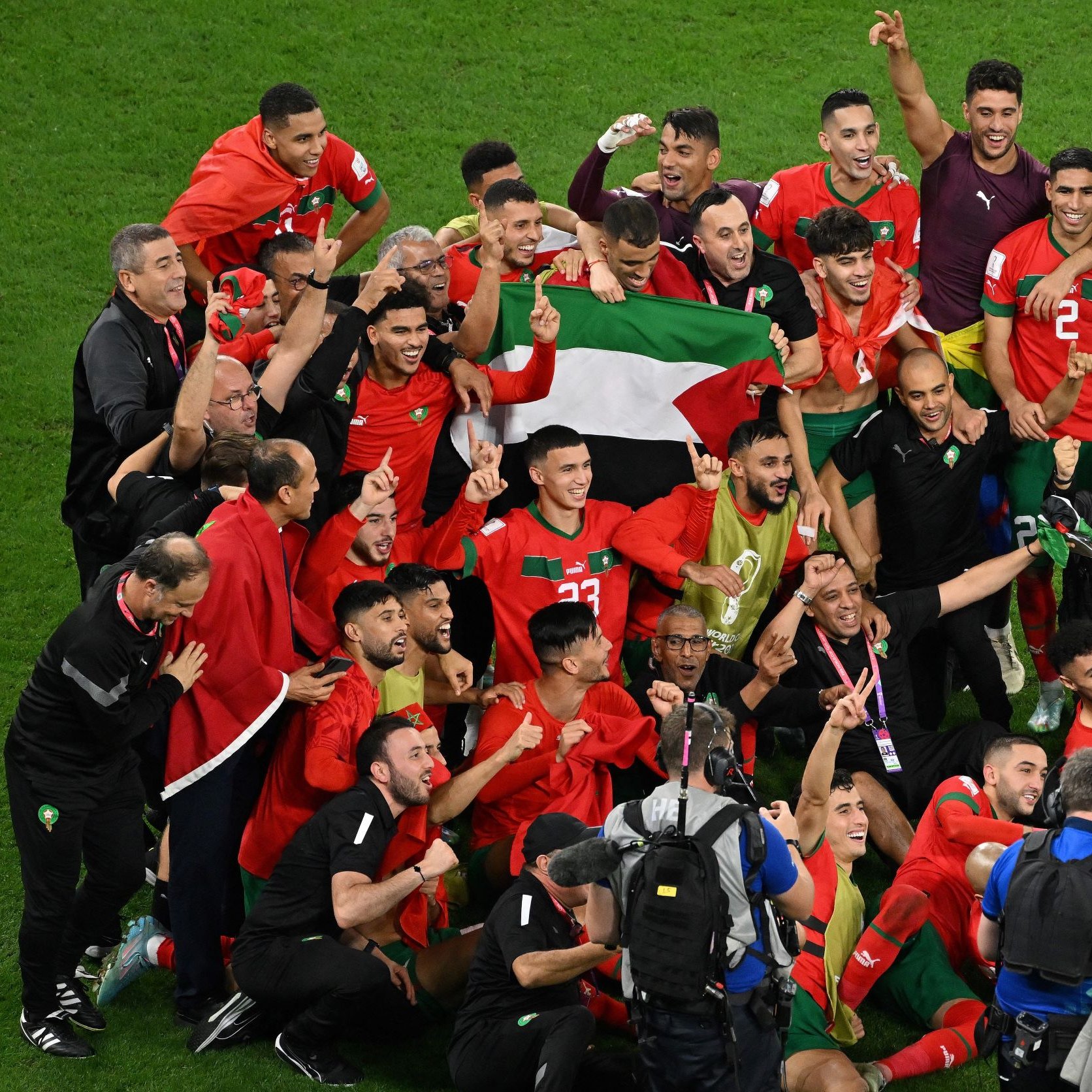 این تصویر نشان‌دهنده خوشحالی و شادی تیم ملی فوتبال مراکش پس از یک پیروزی بزرگ است. بازیکنان و اعضای کادر فنی تیم در حال جشن گرفتن هستند و بعضی از آن‌ها پرچم مراکش را در دستان خود دارند. فضای تصویر پر از انرژی مثبت و شادی است و بازیکنان با لبخند و هیجان در کنار یکدیگر قرار دارند. همچنین تعدادی از عکاسان و اعضای رسانه نیز در حال تصویربرداری از این لحظه مهم هستند.